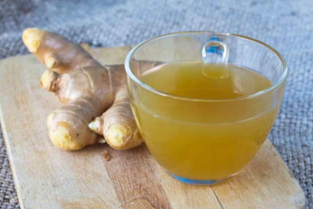 A cup of ginger tea in a transparent glass cup placed on a wooden surface, with fresh ginger root lying beside it.