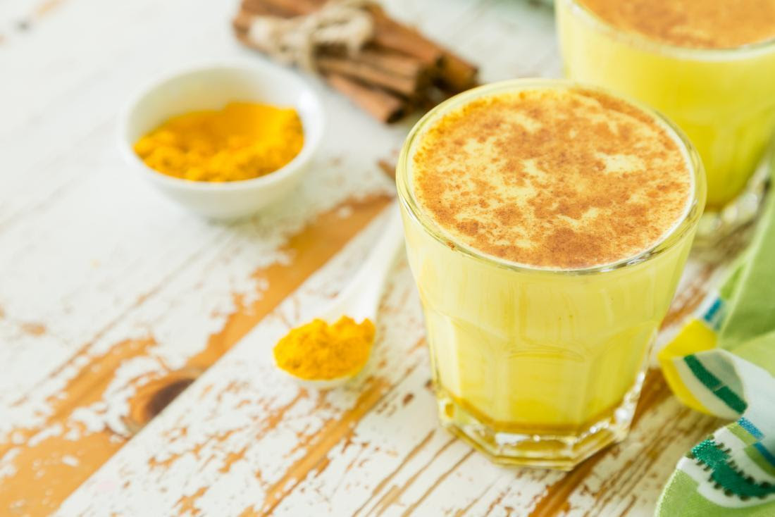 A steaming mug of golden milk topped with a sprinkle of cinnamon, surrounded by turmeric roots and cinnamon sticks on a rustic wooden table.