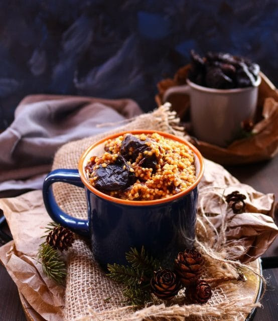 A warm bowl of cinnamon rye berry porridge, topped with chopped walnuts, plump prunes, a dollop of ghee, and a dusting of cinnamon, creating a cozy breakfast scene.