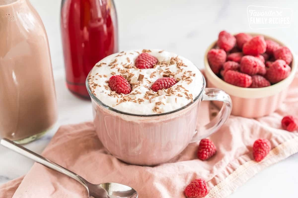 A steaming mug of rich raspberry hot chocolate topped with fresh raspberries and chocolate shavings, set against a cozy winter backdrop with warm lighting.